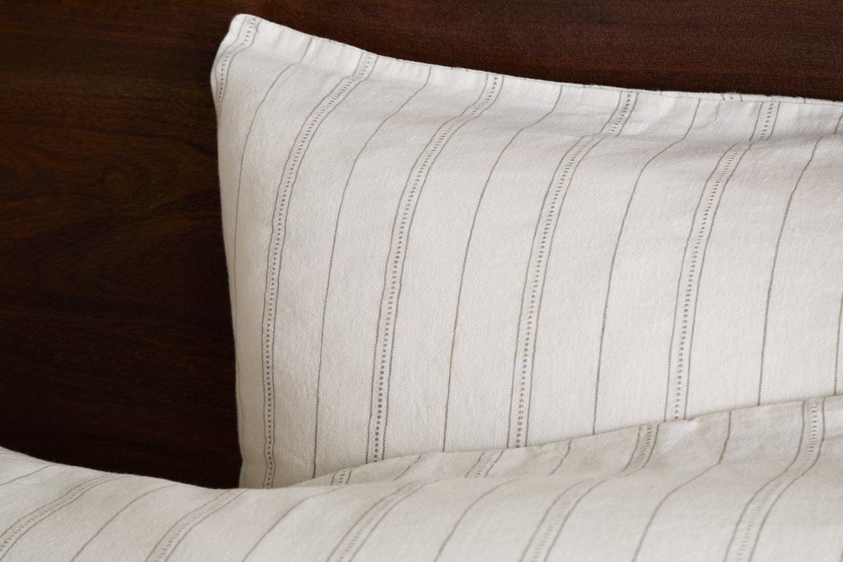 A close-up of a bed with white, thinly striped gray bedding and nkukus Anta Hemp & Cotton Housewife Pillowcases in sage green, set against a dark wooden headboard.