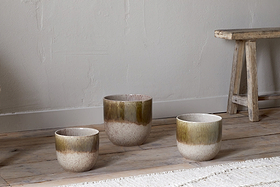 Three Almeida Reactive Glaze Plant Pots by nkuku in Olive & Natural, each a different size with an earthy brown-green to beige gradient glaze, are arranged on a wooden floor beside a rustic bench and a woven rug against a textured wall.