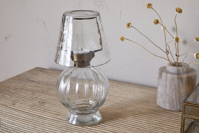 The nkuku Adesola Glass Tealight Holder Lamp in clear, featuring a round base and matching shade, is placed on a striped beige surface beside a vase of dried yellow flowers against a light wall.