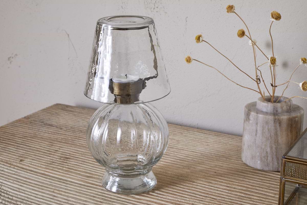 The nkuku Adesola Glass Tealight Holder Lamp in clear, featuring a round base and matching shade, is placed on a striped beige surface beside a vase of dried yellow flowers against a light wall.