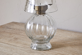 The Adesola Glass Tealight Holder Lamp in clear by nkuku, featuring a round base and metal fixture, sits on a light-textured wooden surface against a plain wall.