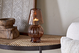 The nkuku Adesola Glass Tealight Holder Lamp in amber sits on a wooden table near a textured artwork and rustic box, casting warm, non-electric light, with part of a pale sofa visible to the right.