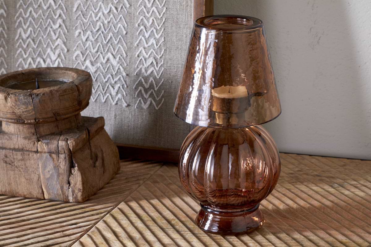 The nkuku Adesola Glass Tealight Holder Lamp in amber, featuring a ribbed base and matching glass shade, rests on wood. Behind it are rustic, square tealight holders and a beige fabric panel with a white zigzag pattern.