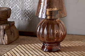 The nkuku Adesola Glass Tealight Holder Lamp in amber, featuring a small rounded shape and ribbed design, sits unlit on a diagonally grooved wooden surface beside rustic wood and white zigzag-embroidered fabric.