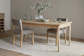 A minimalist dining room featuring the nkuku Anbu Mango Dining Table - Natural, two gray upholstered chairs, a textured rug, and a vase with green branches. Glassware and a bowl sit atop the elegant Anbu collection table.