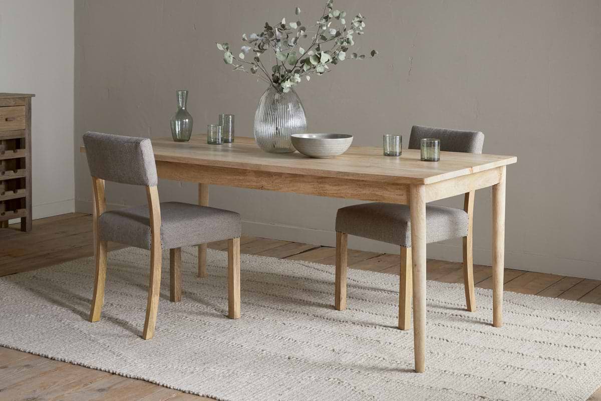 A minimalist dining room featuring the nkuku Anbu Mango Dining Table - Natural, two gray upholstered chairs, a textured rug, and a vase with green branches. Glassware and a bowl sit atop the elegant Anbu collection table.
