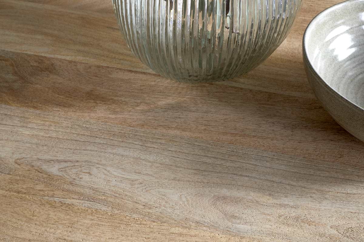 A close-up of the Anbu Mango Dining Table - Natural by nkuku, featuring a light wooden surface with a ribbed transparent glass vase and ceramic bowl partially visible in the top right corner.
