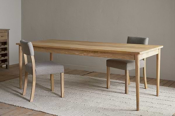 The Anbu Mango Dining Table - Natural by nkuku stands on a textured beige rug, paired with two gray upholstered chairs in a minimalist room featuring wooden floors and a plain light-colored wall.