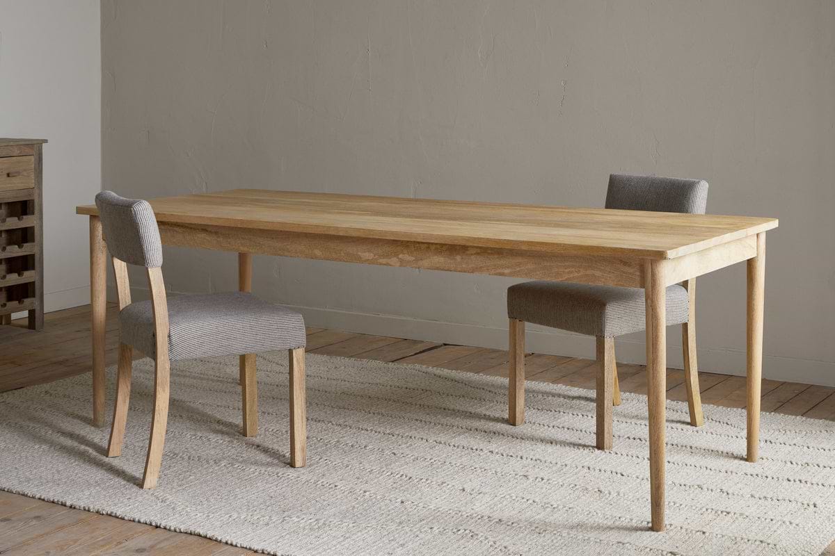The Anbu Mango Dining Table - Natural by nkuku stands on a textured beige rug, paired with two gray upholstered chairs in a minimalist room featuring wooden floors and a plain light-colored wall.