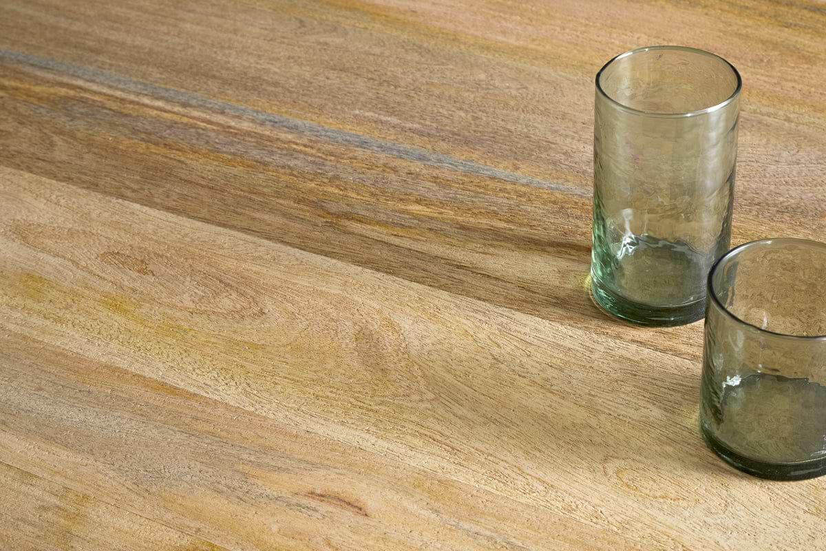 Two empty, transparent glass tumblers rest close together on the lower right corner of an nkuku Anbu Mango Dining Table in natural finish, showcasing the elegant wood grain and creating a refined dining atmosphere.