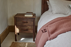The nkuku Anbu Acacia Bedside Table in Washed Walnut, with two drawers, stands by the bed for storage. Three books rest on top. The bed has white and beige striped sheets and a rust blanket, as sunlight streams onto the white floor.