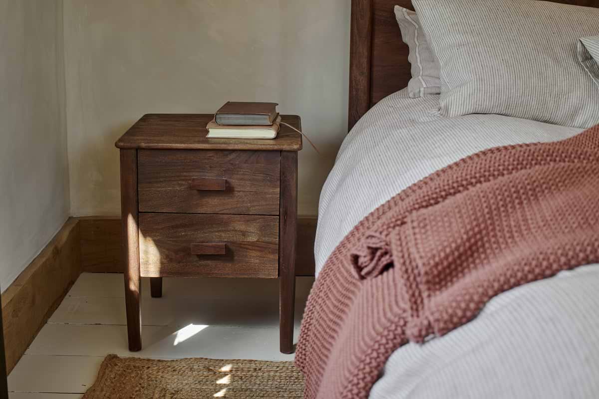 The nkuku Anbu Acacia Bedside Table in Washed Walnut, with two drawers, stands by the bed for storage. Three books rest on top. The bed has white and beige striped sheets and a rust blanket, as sunlight streams onto the white floor.