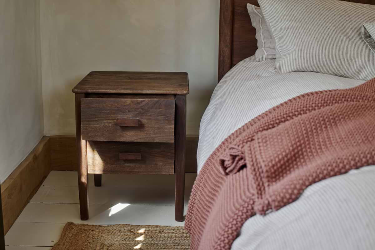 The nkuku Anbu Acacia Bedside Table in Washed Walnut, featuring two drawers, stands by the bed with striped bedding and a dusky pink blanket, offering extra storage and warmth to this softly lit, cozy bedroom.