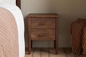 The nkuku Anbu Acacia Bedside Table in Washed Walnut, featuring two drawers, sits between a bed with a pink textured blanket and the wall on wooden flooring. Another textured blanket is partly visible to the right.