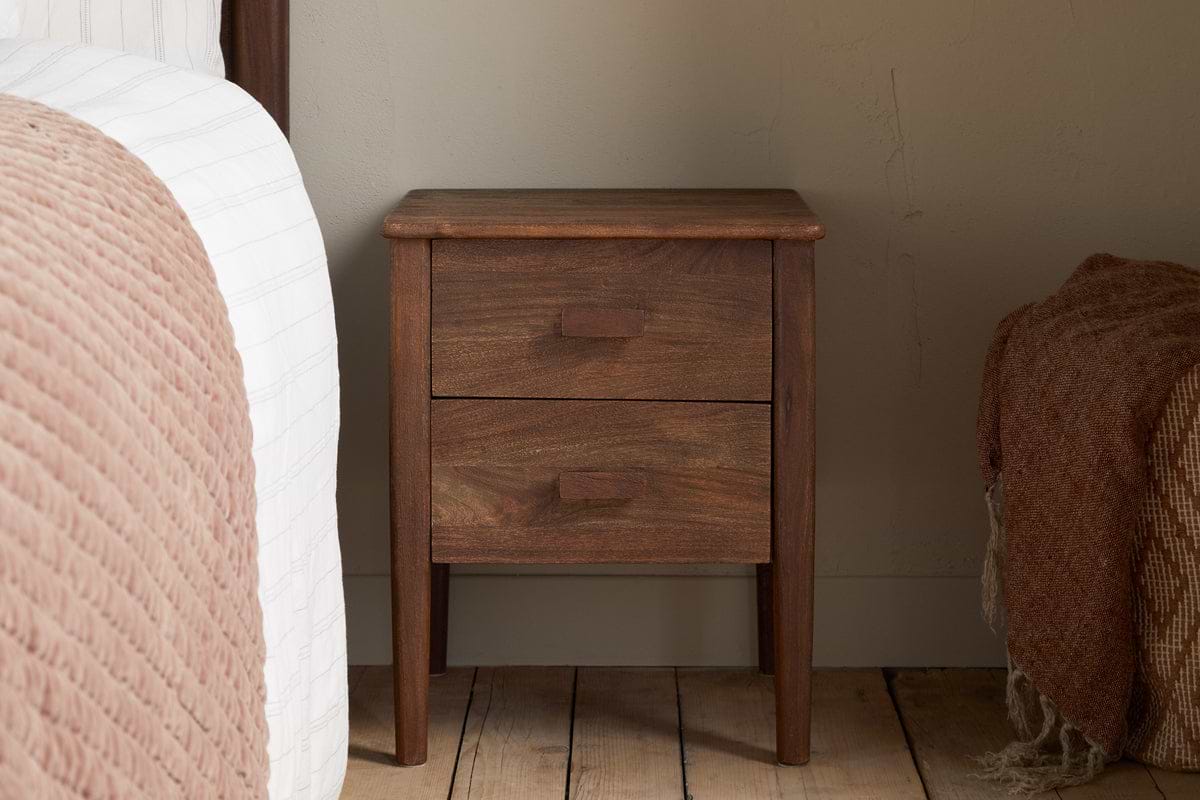 The nkuku Anbu Acacia Bedside Table in Washed Walnut, featuring two drawers, sits between a bed with a pink textured blanket and the wall on wooden flooring. Another textured blanket is partly visible to the right.