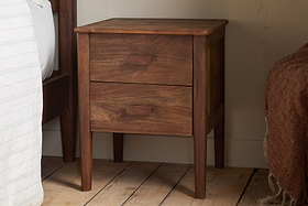 The nkuku Anbu Acacia Bedside Table - Washed Walnut features two drawers with rectangular handles and sits on a wooden floor beside a bed with white striped and brown textured blankets, adding stylish storage to your bedroom.
