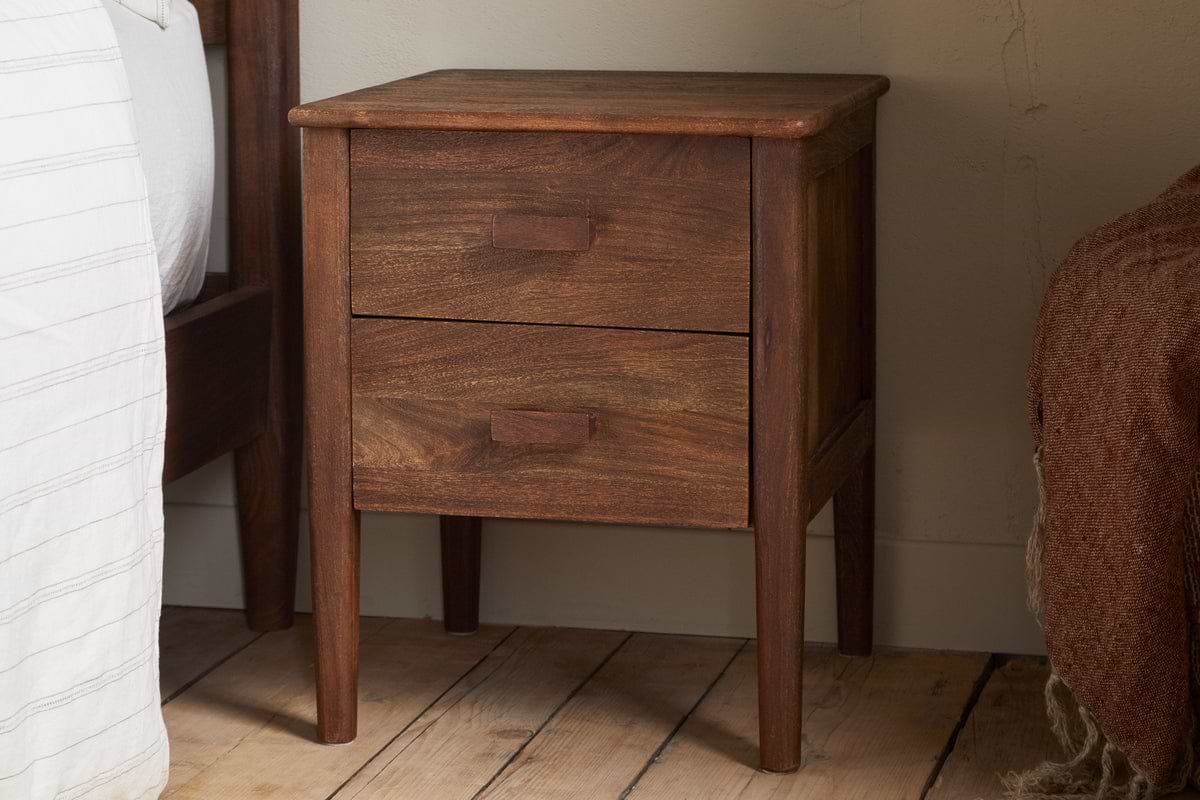 The nkuku Anbu Acacia Bedside Table - Washed Walnut features two drawers with rectangular handles and sits on a wooden floor beside a bed with white striped and brown textured blankets, adding stylish storage to your bedroom.
