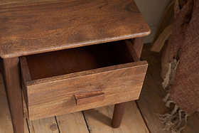 The nkuku Anbu Acacia Bedside Table in Washed Walnut stands on a wooden floor, its drawer partly open. Nearby, a brown tasseled, textured blanket adds warmth and storage to the bedroom.
