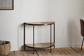 The nkuku Aasha Mango Wood Half Moon Console Table - Natural, with a metal frame and mango wood shelves, stands against a light textured wall on a wooden floor, flanked by a woven basket and part of a brown chair.