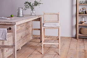 A rustic kitchen features nkuku's Aamani Mango Wood & Cord Counter Chair in natural by a wooden counter holding a pitcher, glass, vase with greenery, and lime. Shelves above display jars, baskets, and covered dishes.