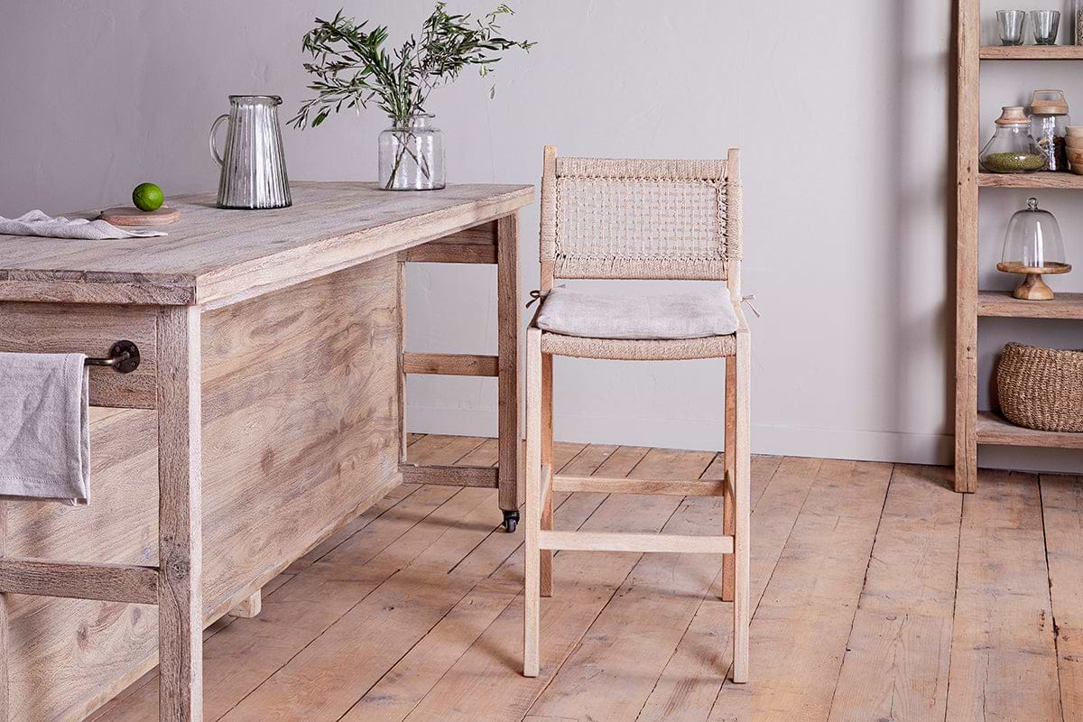 A rustic kitchen features nkuku's Aamani Mango Wood & Cord Counter Chair in natural by a wooden counter holding a pitcher, glass, vase with greenery, and lime. Shelves above display jars, baskets, and covered dishes.