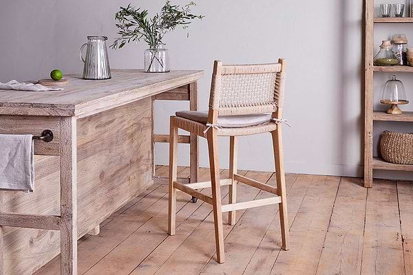 An Aamani Mango Wood & Cord Counter Chair by nkuku sits at a rustic kitchen island topped with a glass jug, lime, and a small vase of greenery. Shelves in the background display jars, baskets, and dishes for added charm.