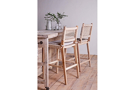 Two nkuku Aamani Mango Wood & Cord Counter Chairs in natural sit by a light wood counter topped with glassware, plates, and a vase of green branches. A wooden floor and plain light wall complete the serene setting.