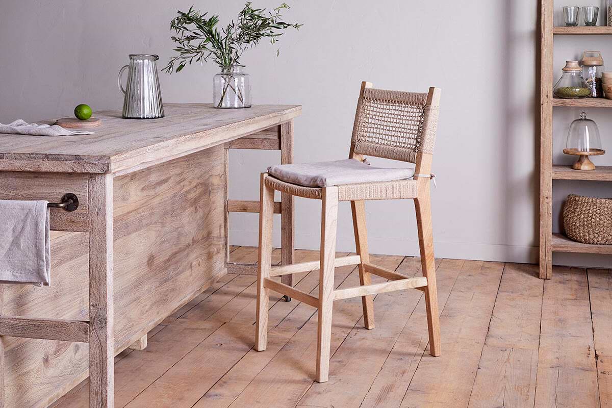 A rustic kitchen features the nkuku Aamani Mango Wood & Cord Counter Chair – Natural, showcasing a recycled paper cord woven back and cushioned seat beside a wooden island with a glass pitcher, vase of greenery, lime, shelving, and baskets in the background.