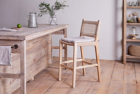 A rustic kitchen features the nkuku Aamani Mango Wood & Cord Counter Chair – Natural, showcasing a recycled paper cord woven back and cushioned seat beside a wooden island with a glass pitcher, vase of greenery, lime, shelving, and baskets in the background.