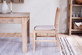 The Aamani Mango Wood & Cord Dining Chair by nkuku, crafted from natural mango wood with a beige cushion, pairs with a matching table. A woven rug, shelf of arranged dishes, and soft lighting complete the rustic, neutral-toned space.