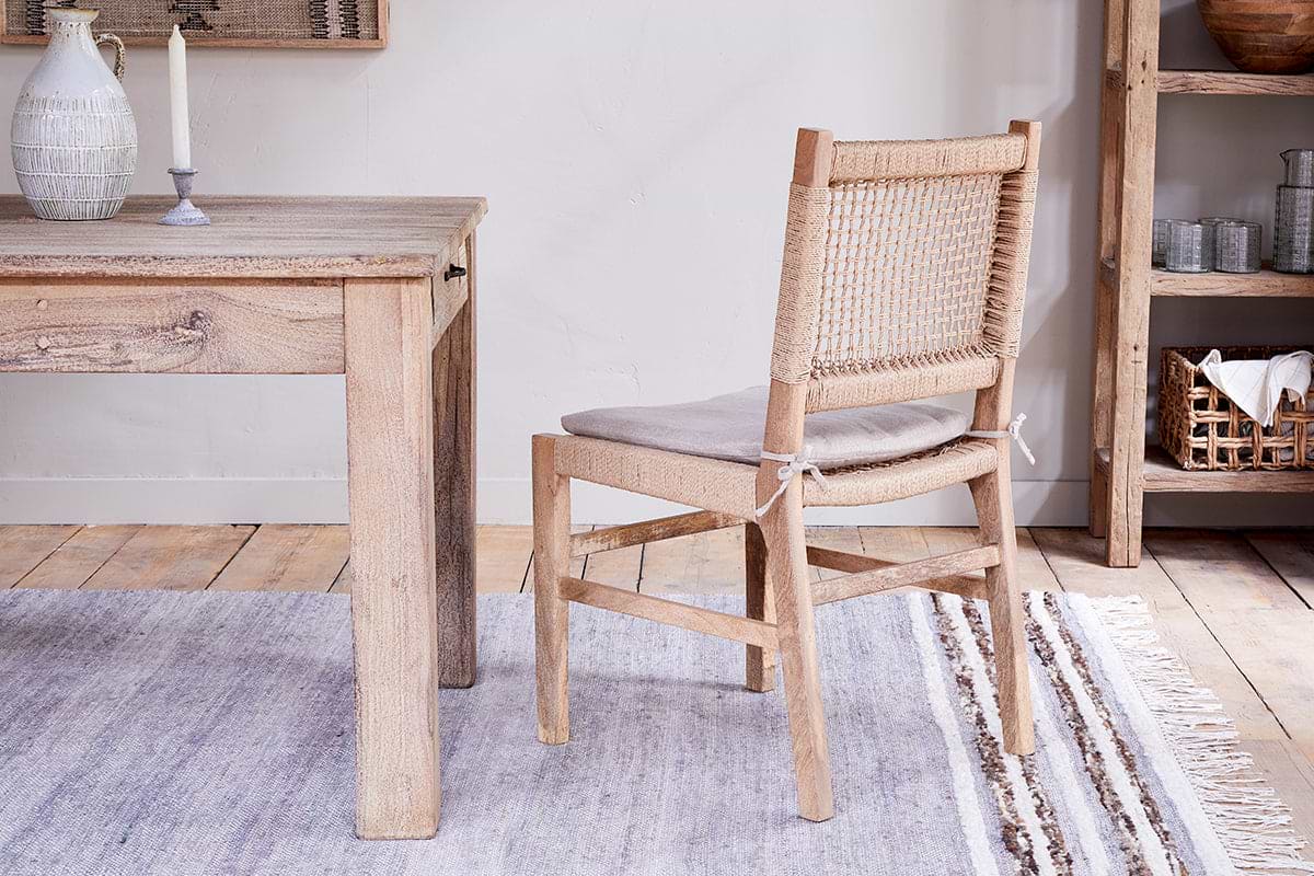 The nkuku Aamani Mango Wood & Cord Dining Chair in natural sits next to a reclaimed wood table, styled on a grey rug with a white candle, ceramic vase, and wooden shelving for a neutral boho look.