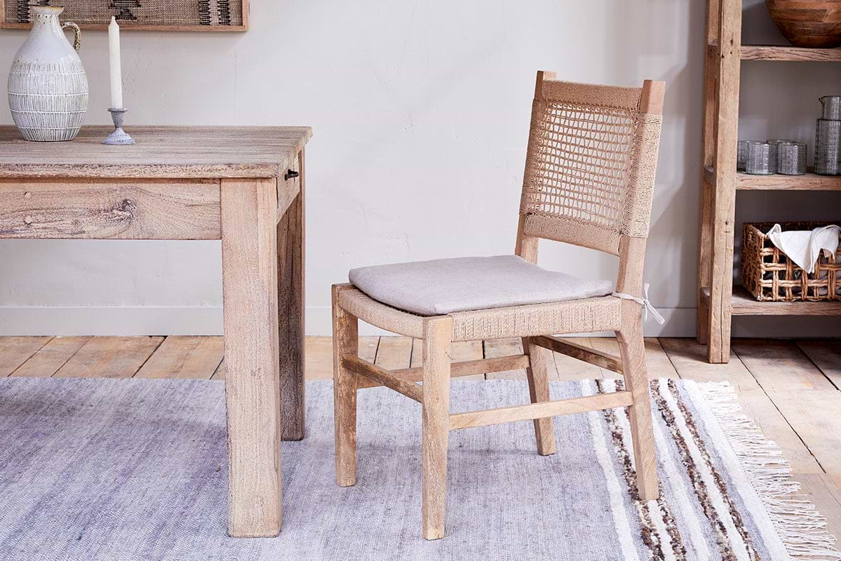 A light wood dining table with a carved pitcher & candle sits next to the nkuku Aamani Mango Wood & Cord Dining Chair - Natural, featuring a light grey cushion, on a striped rug. A rustic wooden shelving unit is on the right.