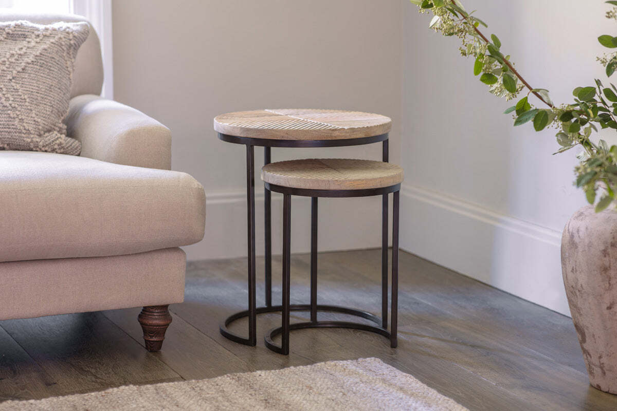 The nkuku Aasha Carved Mango Wood & Iron Nesting Side Tables (Set of 2, Natural) sit beside a neutral-toned sofa, featuring hand-carved mango wood tops and black metal frames.