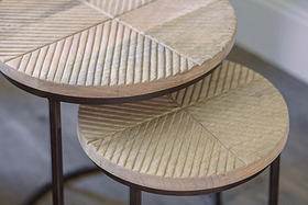 Close-up of nkuku’s Aasha Carved Mango Wood & Iron Nesting Side Tables (Set of 2): each round table features hand-carved, geometric mango wood tops with diagonal grooves and thin dark iron legs, shown on a wood floor by a white wall.
