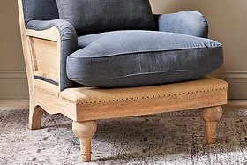 The Abe Deconstructed Linen Armchair by nkuku features a mango wood frame, light wooden legs, beige linen seat base, and thick charcoal cushions. It sits on a patterned rug in a room with neutral walls.