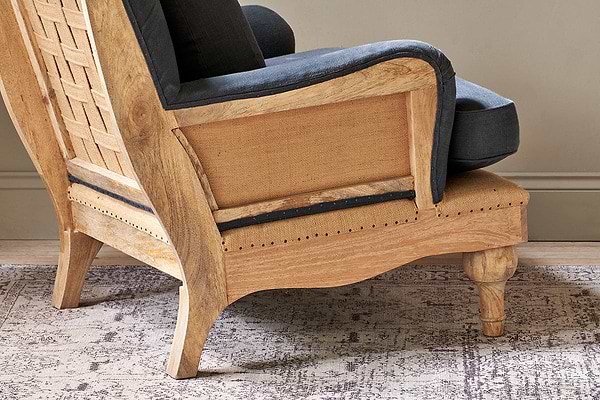 The nkuku Abe Deconstructed Linen Armchair - Charcoal features a light mango wood frame with visible grain, a woven back panel, dark gray linen upholstery, and rests on a patterned rug against a neutral beige wall.