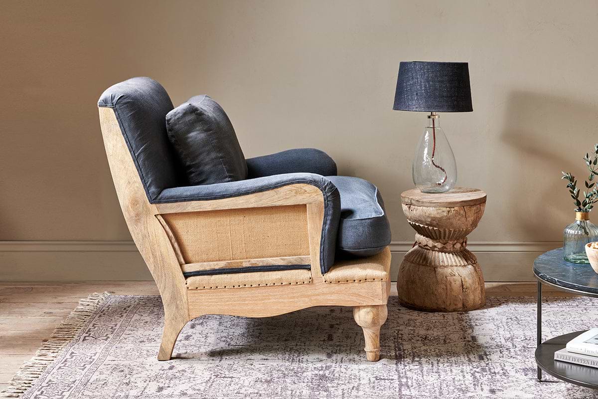 A side view of the nkuku Abe Deconstructed Linen Armchair in charcoal, featuring a mango wood frame, sits beside a carved side table with a clear glass lamp and dark shade on a patterned rug, with a coffee table and plant nearby.