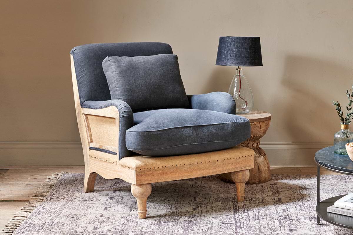 The nkuku Abe Deconstructed Linen Armchair in charcoal sits on a patterned rug. Next to it is a small side table with a lamp and a vase of greenery.