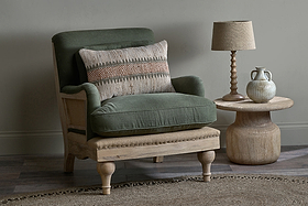 An Abe Deconstructed Linen Armchair - Olive by nkuku, with green cushions, sits beside a round wooden side table holding a beige lamp and ceramic jug. The minimalist space features earthy tones and a woven rug.