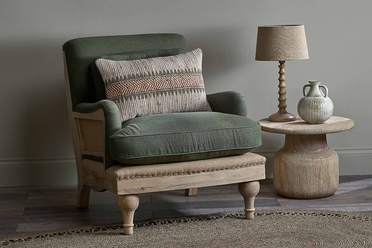 An Abe Deconstructed Linen Armchair - Olive by nkuku, with green cushions, sits beside a round wooden side table holding a beige lamp and ceramic jug. The minimalist space features earthy tones and a woven rug.