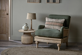 An olive Abe Deconstructed Linen Armchair by nkuku, with a patterned pillow, rests beside a round wooden table holding a lamp and textured vase. A circular jute rug anchors the cozy arrangement.