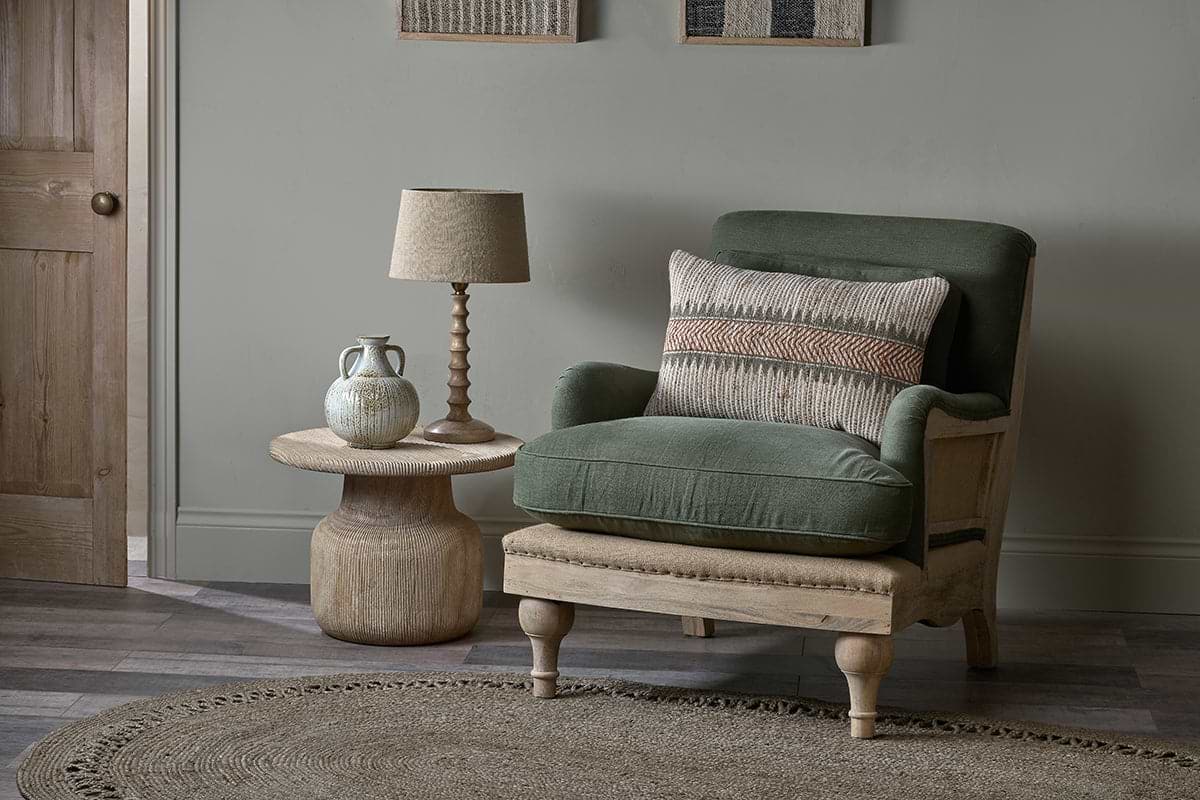 An olive Abe Deconstructed Linen Armchair by nkuku, with a patterned pillow, rests beside a round wooden table holding a lamp and textured vase. A circular jute rug anchors the cozy arrangement.