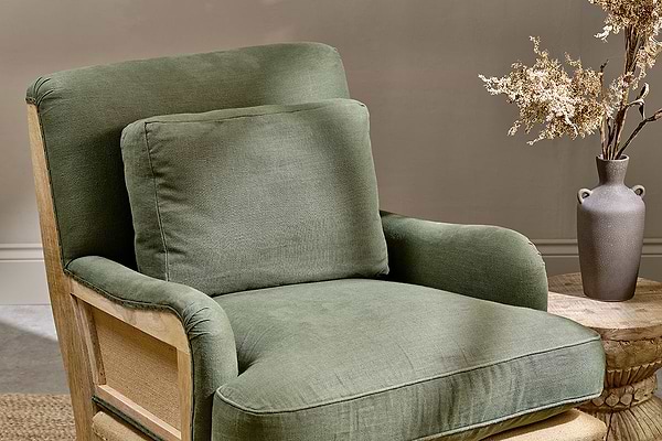 The nkuku Abe Deconstructed Linen Armchair - Olive, paired with a matching cushion, sits beside a round wooden side table topped with a gray ceramic vase and dried branches. A light beige wall highlights the chair’s wooden armrests.
