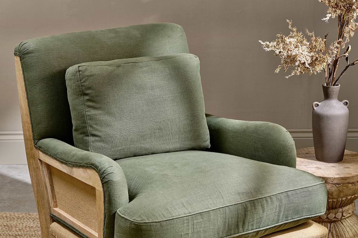 The nkuku Abe Deconstructed Linen Armchair - Olive, paired with a matching cushion, sits beside a round wooden side table topped with a gray ceramic vase and dried branches. A light beige wall highlights the chair’s wooden armrests.