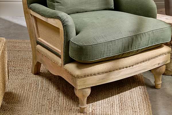 The nkuku Abe Deconstructed Linen Armchair in Olive features green linen cushions, a woven cane side panel, a rustic wood frame, burlap seat, and turned wooden legs, all displayed atop a jute rug.