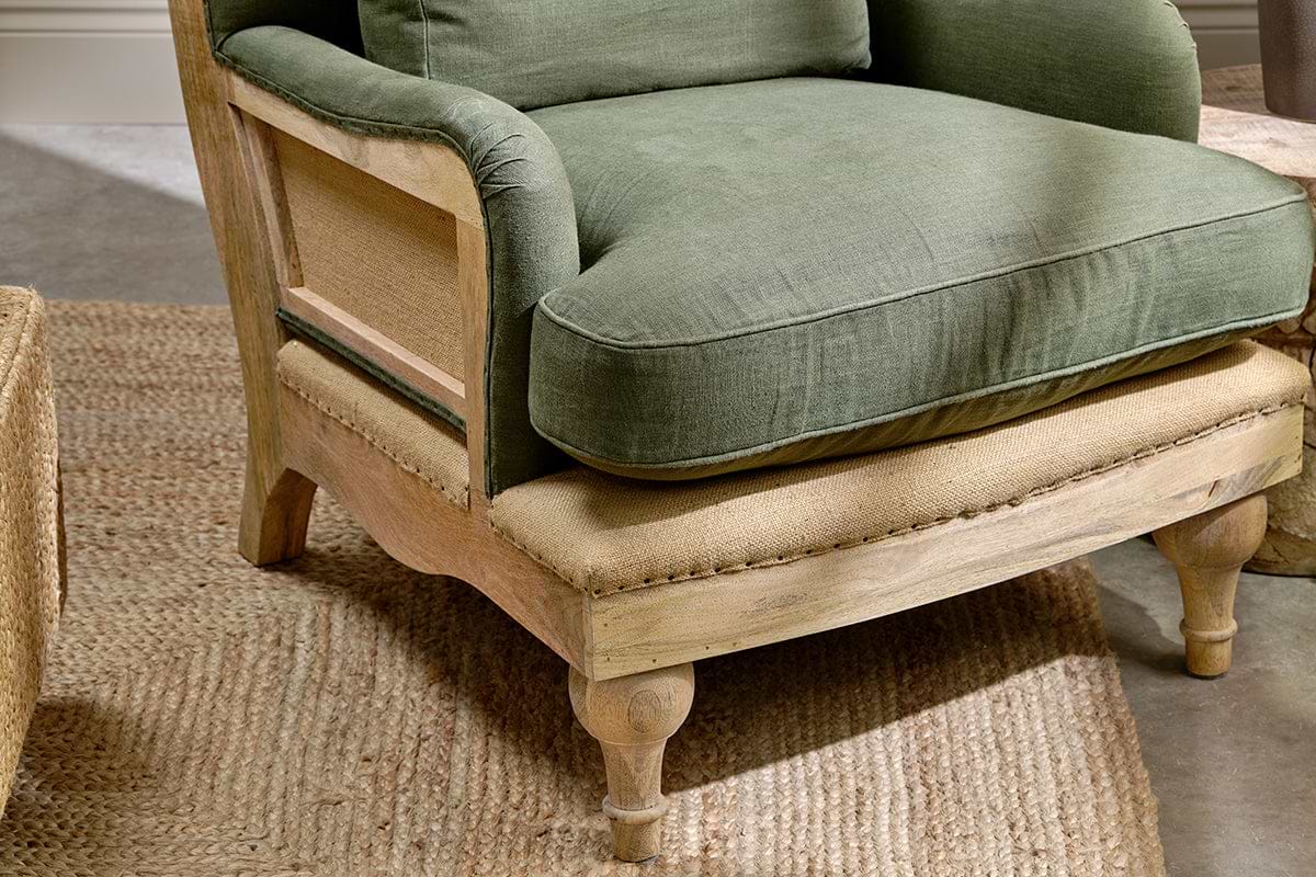 The nkuku Abe Deconstructed Linen Armchair in Olive features green linen cushions, a woven cane side panel, a rustic wood frame, burlap seat, and turned wooden legs, all displayed atop a jute rug.