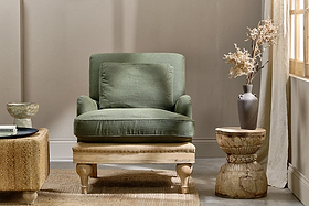 A cozy living room showcases the nkuku Abe Deconstructed Linen Armchair in olive, complete with matching cushion and wooden frame. Nearby, a rustic wood side table holds a gray vase of dried flowers. Sunlight pours through beige curtains.