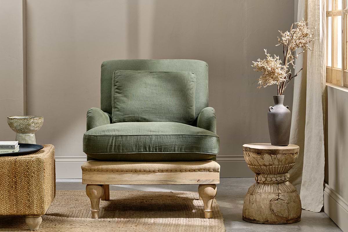 A cozy living room showcases the nkuku Abe Deconstructed Linen Armchair in olive, complete with matching cushion and wooden frame. Nearby, a rustic wood side table holds a gray vase of dried flowers. Sunlight pours through beige curtains.