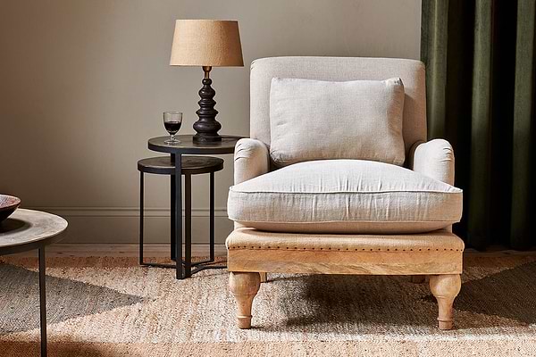 The Abe Deconstructed Linen Armchair - Stone by nkuku sits atop a woven rug. Next to it, a round black side table holds a tan-shaded lamp and glass of red wine, all set against green curtains for an inviting atmosphere.