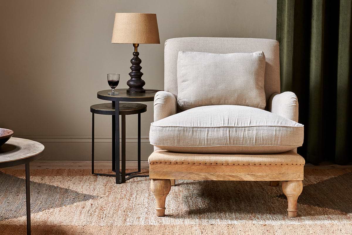 The Abe Deconstructed Linen Armchair - Stone by nkuku sits atop a woven rug. Next to it, a round black side table holds a tan-shaded lamp and glass of red wine, all set against green curtains for an inviting atmosphere.